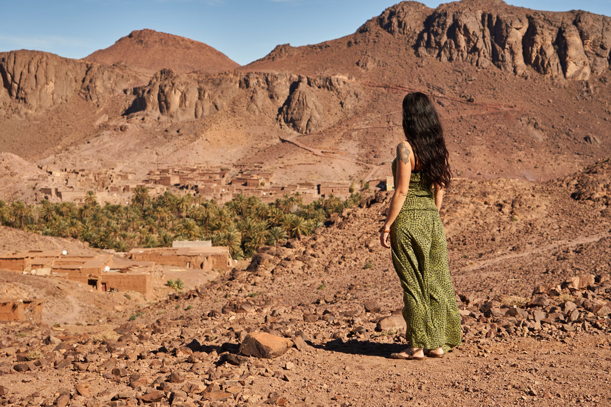Back view of young brunette lady standing between desert lands near ancient constructions and hills in Marrakesh, Morocco